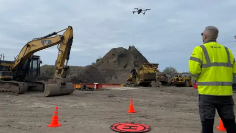 Environment Agency A man in a high-visibility yellow jacket flies a drone above a large mound of waste.