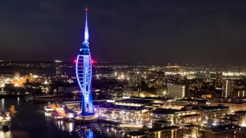 Getty Images View across Portsmouth city at night. Buildings are lit up with lights whilst the sky is dark. In the foreground is the Spinnaker Tower, an iconic tall structure in the shape of a ship's sale which is illuminated blue.
