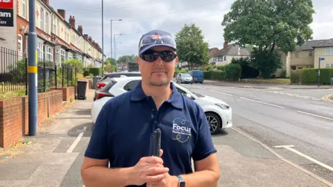 A 57-year-old man, Rob, stands in the middle of a pavement, with cars behind him. He wears a blue polo-shirt and black sunglasses. He holds his mobility cane in front of him.