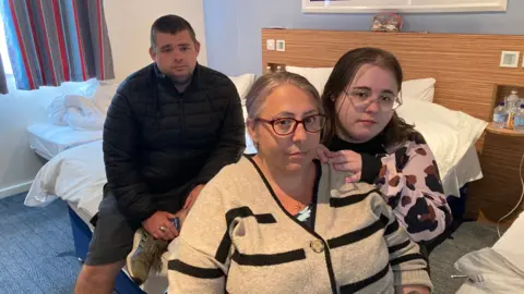 A man and two women sat on a bed in a hotel room. Both women are wearing glasses and all three are looking towards the camera. There is a window and blue and red curtains in the background.