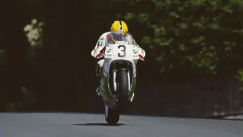 Getty Images Joey Dunlop is racing a white and green bike with the number three in black on the front. The bike is on its back wheel as he races along the road. He is wearing a yellow helmet, a white racing suit and red gloves. Behind him is the darkness of the green trees.