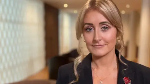 Katie Amess is wearing a black suit jacket with a poppy brooch. She has blonde hair, a crucifix pendant on a chain and is stood in a hall with pillars  