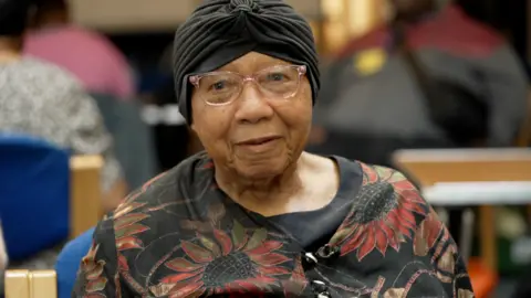 A black lady with a hair wrap and top with red flowers on black smiles into the camera