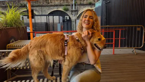 Jenn Nimmo-Smith - a woman with long blonde hair - sits on a bench in a bar's outdoor area and cuddles her dog Scout, a large golden retriever