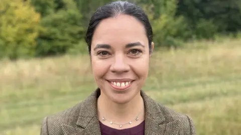 Marnie Lovejoy with dark hair in a ponytail wearing a tweed jacket and purple round neck blouse