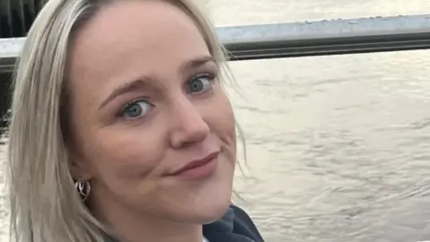 Chloe Wilson stands in front of a river. She has long blond hair and blue eyes, her face is close to the camera and she is wearing a nose ring and earrings. The collar of her grey jacket can also be seen