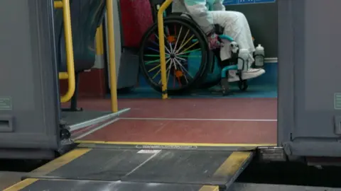 A wheelchair user is inside a bus on the wheelchair space, positioned near the door. A black and yellow ramp extends from the bus doorway to the ground for accessibility.