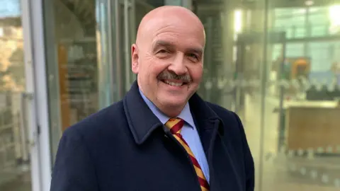 BBC A man looking to his right side smiling at the camera. He is wearing a navy blue coat jacket on top of a light blue shirt and a red and yellow striped tie. Behind him is the blurred glass door of a government building.