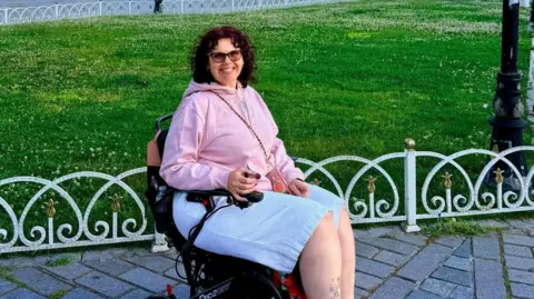 A woman with dark curly hair is sitting in a wheelchair and is wearing a pink hoodie and white shorts. She is in the middle of a park and smiling.