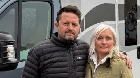 Adrian and Joanne Fenton wearing coats and standing in front of their navy and white motorhome, which is parked in front of their house. They have serious expressions on their faces.