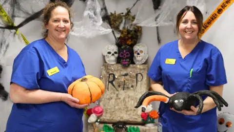 NHS Somerset Foundation Trust Two women in scrubs are holding up Halloween decorations, standing in front of a display including a grave and skulls.