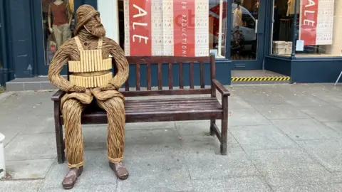 A wicker figure depicting a bearded RNLI crewman from 1886 - including a helmet and lifejacket - is sitting on a bench outside a shop in a town centre. The figure has its hands resting on its legs and is looking to its left.