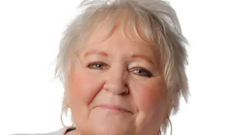 Anita Harrison. She has short white hair and is wearing hooped earrings