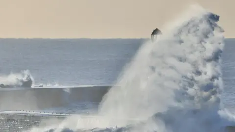 Dan Seaham Harbour Lad A large wave which is almost completely covering the lighthouse. Only the top of it can be seen. There is a pier to the left of the photograph which is being hit by a smaller wave. The sea behind it is a dark blue colour and looks calm. The sky is a beige colour.