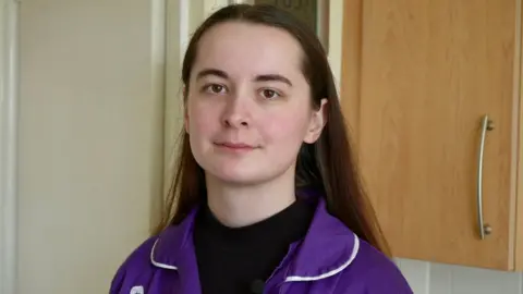 Mollie Cole‑Wilkin looks at the camera. She is wearing a purple uniform with white piping around the edge. She has a black turtle neck top on underneath it. Mollie is standing in the kitchen of Stephen's house. You can see her head and shoulders and there is a wooden kitchen cabinet in the background.