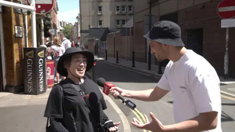 A woman on the street laughing as a reporter holds a microphone to her mouth. She is wearing a black shirt and a black brimmed hat. the reporter also as a black bucket hat and a white t-shirt. They are on the street in Cardiff. 