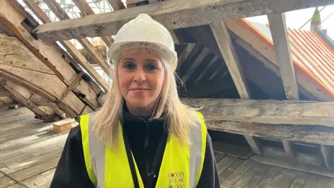 Photograph of Jany Baxter from Stockport Council. She is pictured inside a loft which is undergoing restoration work. She wears a yellow hi-vis on top of black jacket. She has long blonde hair under a white hard hat.