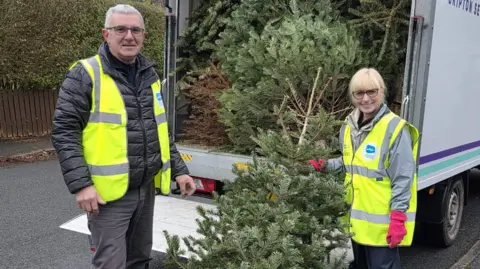 A man and woman in hi-vis vests loading a green fir tree into the back of a van.