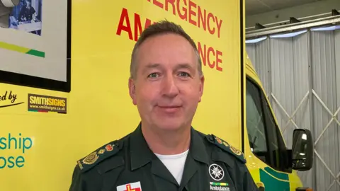 BBC A man wearing an ambulance uniform standing in front of a yellow and green ambulance inside a garage.