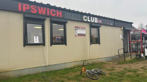 A pale clubhouse building with a black and red "Ipswich BMX club" banner across the top. A BMX bike with a red front lays in front of the clubhouse.