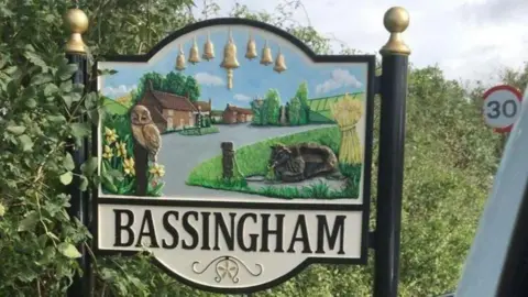 A large village sign on the road into Bassingham. An image depicts houses, a road, a green, a cow, an owl, trees, flowers and symbols of village life, including a sheaf of wheat and church bells. A 30mph road sign can be seen in the background.