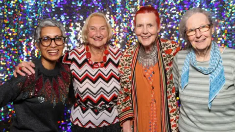 Victoria Dawe Four women, who work as volunteers and supporters of the charity Together Co, standing side by side in front of a shiny and sparkly back drop. 