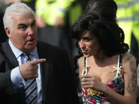 PA Media Man in suit and tie points at woman with big black hair in colourful top smiling