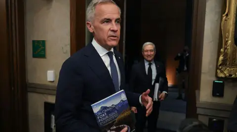 An image of Mark Carney in the foyer of Canada's House of Commons. He is seen speaking to a person off-camera while holding a document titled 'Un Canada Fort Pour Tous'. Beside him is finance minister Francois-Philippe Champagne, who is looking over at Carney and is holding the same document.