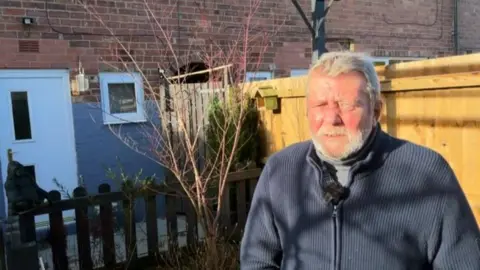 A man with short white hair and a white beard stands in a garden on a sunny day. He is wearing a blue zipped top.