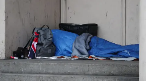 A person covered under a blue sleeping bag with a grey bag next to them. They appear to be lying at the top of stone steps and in front of a wall.