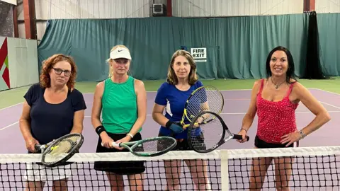Four women standing at the net of the Withdean indoor courts. They are all holding tennis rackets  and are wearing sports gear.