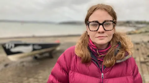 Sophie Pierce wears a dark pink puffy jacket and a stripy scarf of the same colour. She has thick, square framed glasses and her hair is loose around her shoulders. In the background there is open water with a longboat on the shore. 