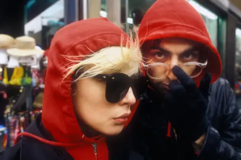 Martyn Goddard Debbie Harry and a Chris Stein wear red hooded jackets and dark sunglasses on a New York street, with Harry in profile and the other figure adjusting his glasses while looking at the camera.