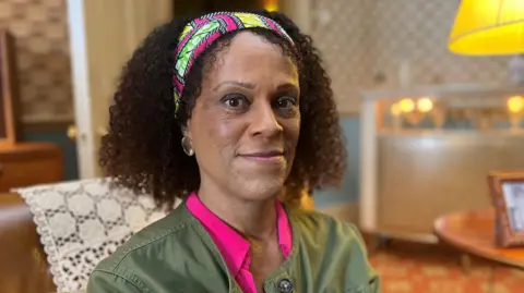 Bernadine Evaristo wearing a green jacket, pink shirt and colourful headband sitting on a brown sofa