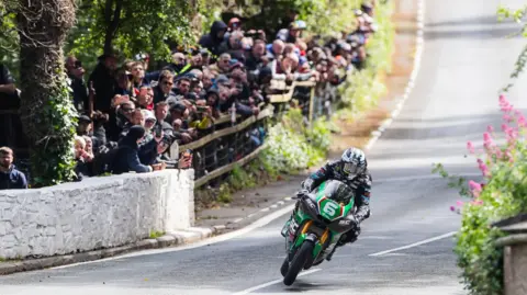 ISLE OF MAN TT Spectators watch on from behind a fence at the bottom of a dip as Michael Dunlop, on a green and black bike with the number six on the front, races past. 