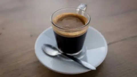 Transparent mug of expresso and a teaspoon on a white saucer.