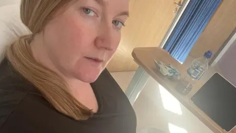 Hayley Burrows / Brain Tumour Research Hayley sits on a hospital bed wearing a black scoop-neck t-shirt