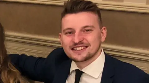 Family A smiling man with brown hair and a small beard wearing a dark suit, white shirt and dark tie