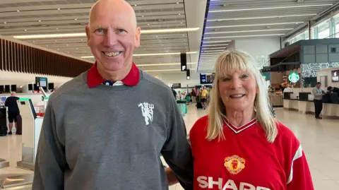 Jim is bald and is wearing a grey crew neck sweater with a white Manchester United crest while his wife has blonde hair and is wearing a replica Manchester United shirt