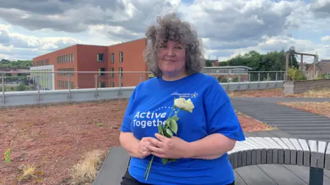 A woman with grey curly hair is sitting outdoors on a bench and smiling. She is wearing a bright blue t-shirt with the words Active Together and is holding a white rose