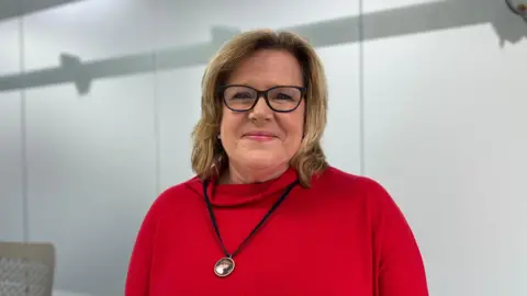 Rachel Lucas is wearing a red roll-neck jumper and a black necklace. She is sitting in a grey and white office.