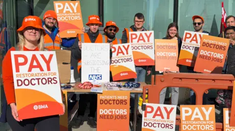 About a dozen banners, with some including the words pay restoration for doctors, are either being held by people outside a building or are vertical on the ground.