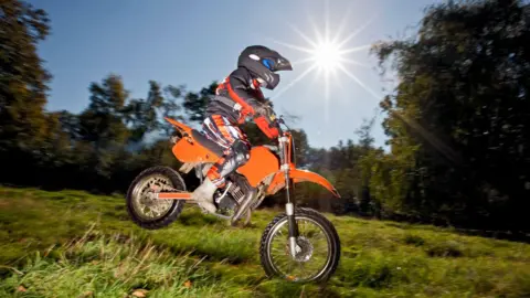 Cavan Images/Getty Images A young boy riding an orange scrambler bike on a grassy area with trees in the background. He is wearing a black helmet, a black and orange biker's suit and rubber boots. 