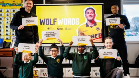 Joao Gomes is dressed in a black and gold Wolves tracksuit, stands behind a row of boys and girls with another male club representative. They are all holding up motivational sentences about speech. There is a poster of Gomes and the words "Wolf Words" behind them all. The pupils are in green and green school uniform. 