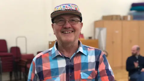 Joe Hoare, a man wearing a red, blue and white checked shirt, glasses and a cap with multicoloured letters on it, smiling for the camera.