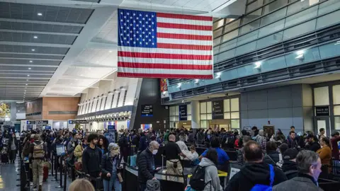Anadolu via Getty Images Long lines of people with suitcases wrap around an airport terminal. An American flag hangs overhead. 