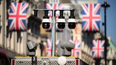 Getty Images Three spherical black cameras are shown, hoisted on a post on top of a police van. In the background is the Union Jack bunting of Regent Street, by Oxford Circus tube station.