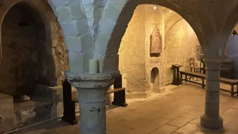 Eastbridge Hospital A Medieval room with low, vaulted ceilings made from stone. Wooden benches and a wall decoration can be seen.