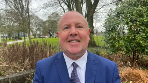 Jim O'Boyle, cabinet member for jobs at Coventry City Concil smiles at the camera. He is wearing a blue suit jacket, white shirt and blue tie. Behind him we see trees, grass and bushes.