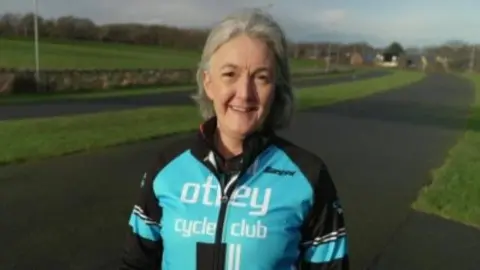 A smiling woman looking straight ahead. She is wearing a black and light blue zip up jacket with the words 'Otley cycle club' on the front. She is stood outside and in the background is a cycle pathway and a grassed area.